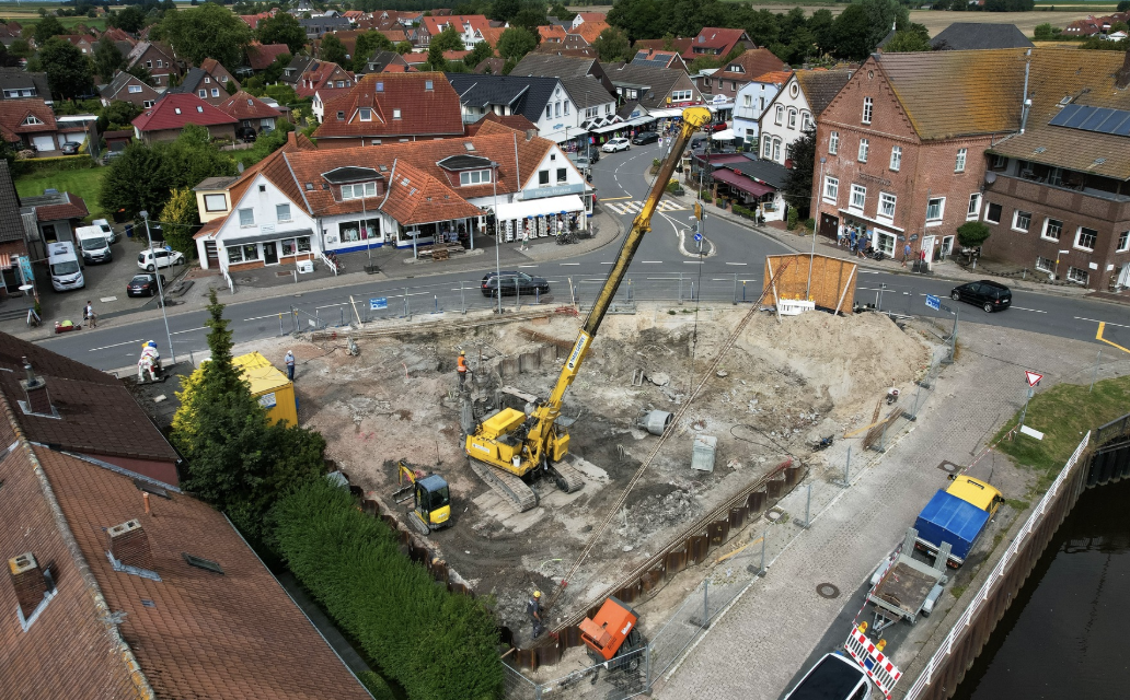 Ein großes gelbes Bohrgerät steht im ausgehobenen Baufeld, umgeben von weiteren Baumaschinen. Rundherum sind Wohn- und Geschäftsgebäude sowie Straßenverkehr zu sehen.